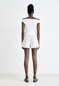 Weißes Off-Shoulder-Oberteil, graue gestreifte Shorts und schwarze Riemchensandalen. Der Schnitt ist lässig, mit einer strukturierten Optik und einem elastischen Taillenbund.