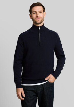 Man met kort bruin haar en lichte baard, die een marineblauwe sweater met rits en een donkere spijkerbroek draagt, staand tegen een effen grijze achtergrond.