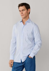 Mężczyzna w jasnoniebieskiej pasiasto-prążkowanej koszuli na guziki z długim rękawem i niebieskich dżinsach, stojący z jedną ręką w kieszeni na tle jednolitej scenerii.