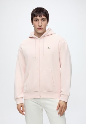 Joven de pie mirando hacia adelante vistiendo una sudadera con capucha rosa claro con cremallera, con un pequeño logo verde de cocodrilo y pantalones blancos.