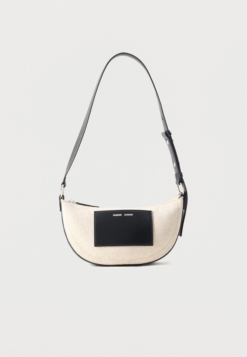 Μπεζ τσάντα ώμου σε σχήμα ημισέληνου με ρυθμιζόμενο μαύρο λουρί και μαύρο ορθογώνιο έμπλαστρο στο μπροστινό μέρος με μικρό λευκό κείμενο.