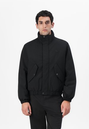Veste noire avec un col haut, une taille élastique et des poches à bouton-pression. Fabriquée en tissu lisse avec un design ajusté et structuré.