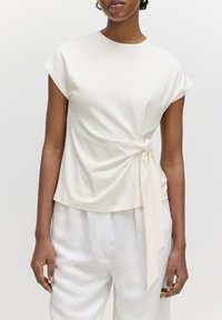 Beige Kurzarmtop mit einem verdrehten Design an der Taille, versehen mit einem gebundenen Akzent, kombiniert mit lockeren weißen Hosen. Glatte Stoffstruktur.