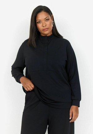 Schwarzer Pullover mit hohem Kragen, langen Ärmeln und einem strukturierten Frontpanel. Weicher Stoff mit lockerer Passform, kombiniert mit passenden schwarzen Unterteilen.