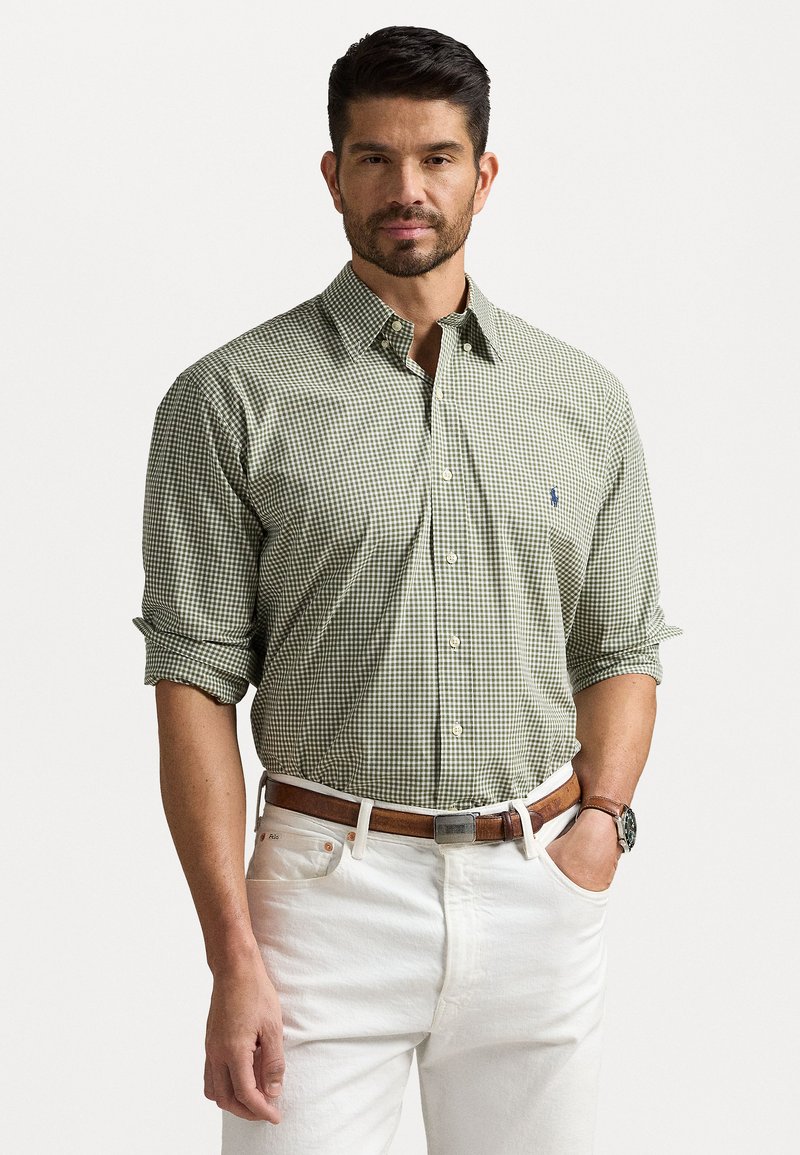 Πράσινο και λευκό καρώ πουκάμισο με γιακά που κουμπώνει, διπλωμένα μανίκια και μικρό μπλε λογότυπο στο στήθος, φορεμένο με λευκά παντελόνα.