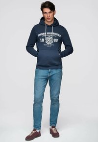 Jeune homme portant un sweat à capuche bleu marine avec un texte blanc et un jean bleu clair, debout devant un fond gris clair uni.