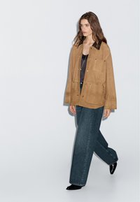 Giacca scamosciata beige con colletto nero, tasche frontali, abbinata a jeans scuri a gamba larga e scarpe nere a punta.