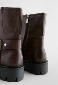 Botas de cuero marrón hasta el tobillo con acabado texturizado, cierre lateral de cremallera y una correa ancha con un acento plateado. Suela de goma negra gruesa.