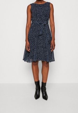 Granatowa sukienka bez rękawów w białe kropki, z paskiem w talii, o długości do kolan, noszona z czarnymi botkami z noskiem w szpic.