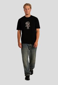 Černé tričko s krátkým rukávem, které na sobě má grafiku detailní lebky ve tvaru panenky. Kombinováno s volnými vybledlými modrými džíny a černou obuví.