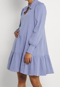 Vaaleansininen, pitkähihainen mekko, jossa on röyhelökaula-aukko ja edessä solmittu vyö, joka sisältää liikkuvan kerroksellisen helman ja teksturoidun kankaan.