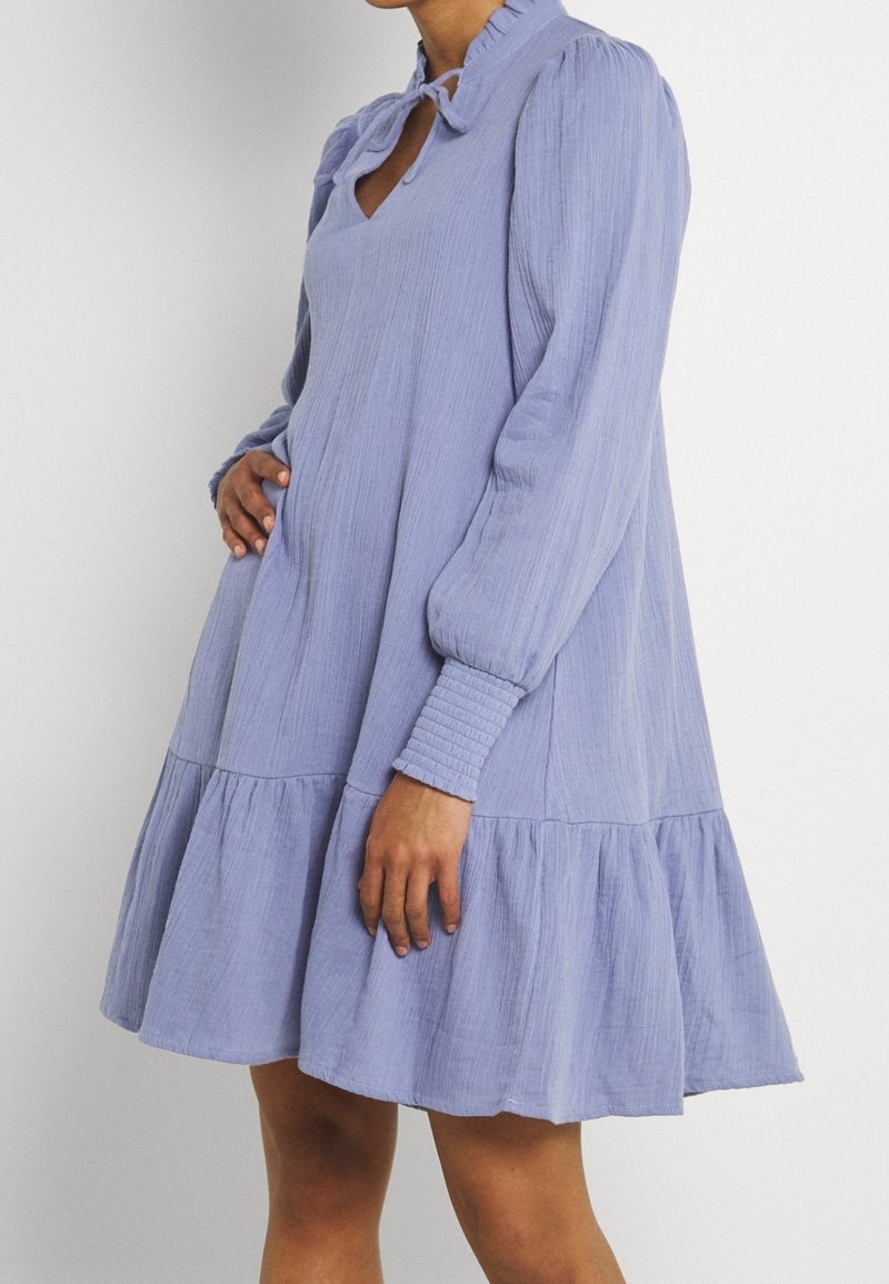 Vaaleansininen, pitkähihainen mekko, jossa on röyhelökaula-aukko ja edessä solmittu vyö, joka sisältää liikkuvan kerroksellisen helman ja teksturoidun kankaan.