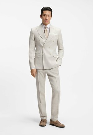 Mężczyzna w beżowym dwurzędowym garniturze, z wzorzystym krawatem i brązowymi mokasynami, stojący z jedną ręką w kieszeni na białym tle.