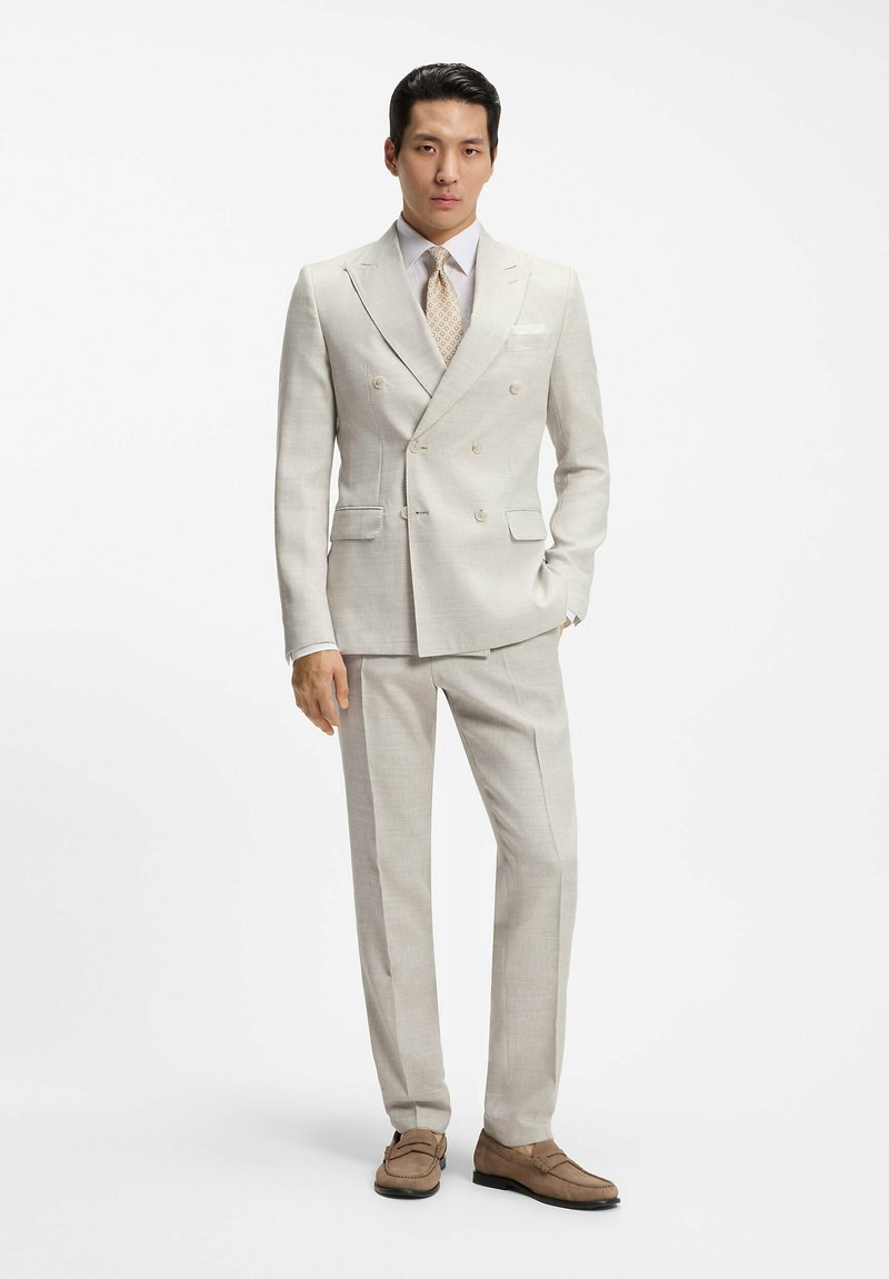 Mand i beige dobbeltradet jakkesæt, mønstret slips og brune loafers, stående med den ene hånd i lommen mod hvid baggrund.