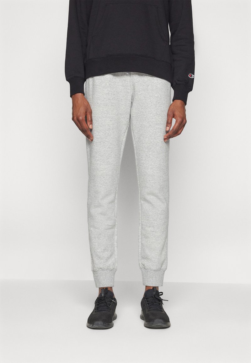 Szürke, szűkített lábú melegítőnadrág, puha anyagból. Gumis derékrész és mandzsetta, fekete kapucnis sweatshirt-tel és fekete sportcipővel párosítva.