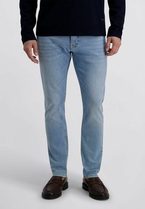 Mann trägt hellblaue, schlanke Jeans mit dunkelbraunen Loafers und einem schwarzen Langarmshirt, steht vor einem einfarbigen Hintergrund.