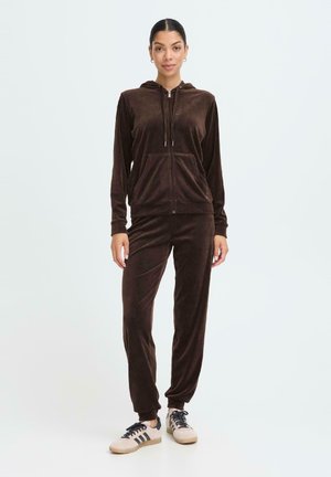 Brown velour tracksuit with zippered hoodie, front pocket, and tapered pants, worn with beige and black sneakers. Smooth texture, casual design.