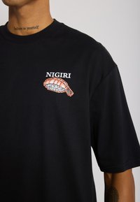 Fekete pamut póló, amelyen egy garnéla nigiri grafikája látható fehér rizzsel, és a "NIGIRI" felirat fölötte. Rövid ujjú; laza fazonú.