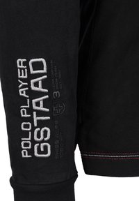 Svart långärmad skjorta med vit och grå broderad text, inklusive "POLOPLAYER GSTAAD" och en schweizisk korsemblem. Detaljer med röd söm.