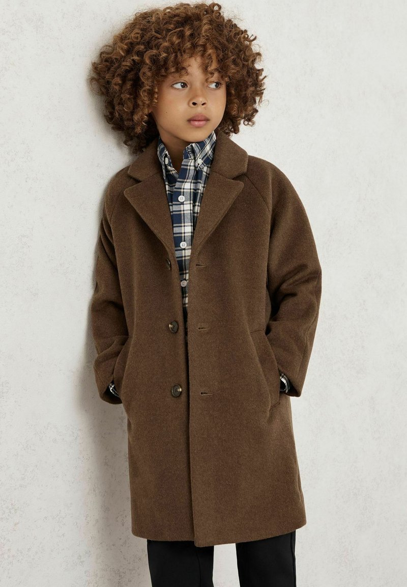 Rudas vilnos paltas su įpjauta apykakle, priekiniais mygtukais ir šoninėmis kišenėmis, uždėtas ant languoto marškinėlių. Kraidė plaukuose, stovi prie sienos.