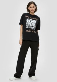 Schwarzes übergroßes T-Shirt mit einem weißen Grafikdesign und Text, kombiniert mit lockeren schwarzen Cargo-Hosen und weißen Sneakern. Einfaches, lässiges Outfit.