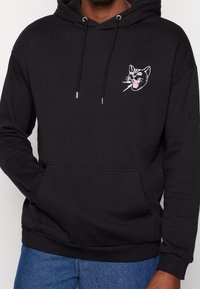Felpa nera in cotone misto, con tasca frontale e design di un volto di gatto ricamato in bianco sul petto. Cappuccio regolabile con lacci.