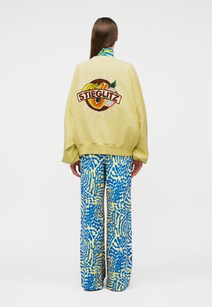 Osoba w nosząc żółtą, oversize'ową kurtkę z owocami i napisem "STIEGLITZ" na plecach, w połączeniu z luźnymi, niebiesko-żółtymi spodniami w wzory.