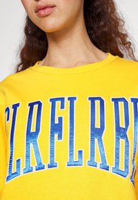 Sudadera amarilla con texto azul en negrita que dice "CLRFLRBB". La tela parece suave, con un diseño clásico de cuello redondo.