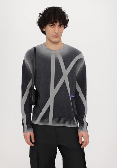 Grauer und schwarzer gestrickter Pullover mit abstraktem Muster und gerippten Bündchen. Dazu eine kleine schwarze Tasche über der Schulter.