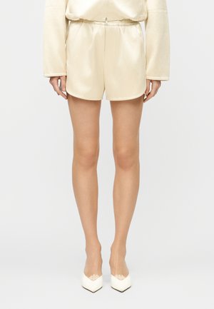 Femme portant un short en satin crème à taille élastique, un haut à manches longues assorti crème et des talons pointus crème avec des détails en dentelle.