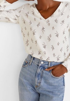 Femme portant un chemisier crème à col en V avec de petits motifs floraux marron et un jean en denim bleu clair taille haute, une main dans la poche.
