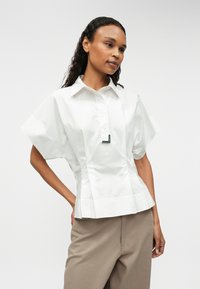 Λευκό μπλουζάκι κοντομάνικο με δομημένη εφαρμογή, κλείσιμο με κουμπί και διακριτική λεπτομέρεια κοντά στο λαιμό. Σχεδιασμένο από απαλό ύφασμα.