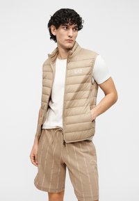 Smėlio spalvos pūstas liemenė su užtrauktuku priekyje ir stendinio tipo apykakle, dėvima ant balto marškinėlių. Derančios smėlio spalvos šortai su logotipo raštu.