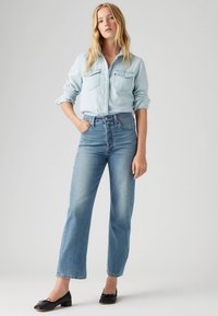 Ljusblå denimskjorta med knappstängning framtill och två bröstfickor, kombinerad med ljusblå högmidjade jeans och svarta ballerina-skor.
