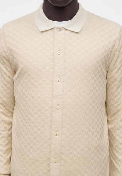 Chemise beige texturée à manches longues avec boutons et col portée sur un sous-vêtement blanc sur une personne à la peau foncée.