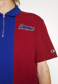 Personne portant un polo zippé bicolore, moitié bleu, moitié bordeaux, avec le logo « Champion » sur la poitrine et la manche, montrant des tatouages sur l'avant-bras.