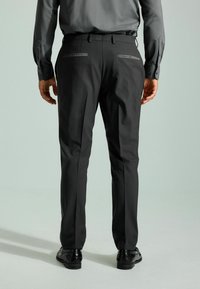 Pantalon noir ajusté, en tissu lisse, coupe slim, deux poches arrière avec un bord epuré, associé à des chaussures noires, vu de dos.