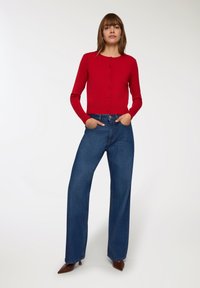 Cardigan rosso aderente con chiusura a bottoni, abbinato a jeans a gamba larga a vita alta blu, completato da décolleté a punta marroni. Sfondo neutro.