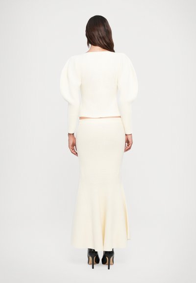 Bela pletena dvodelna obleka, ki vključuje oprijet zgornji del s puff rokavi in dolgo, razširjeno krilo. Nosi se z črnimi škornji z visokimi petami.