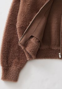 Close-up van een bruine fuzzy jas met een gedeeltelijk open zilveren rits die zachte, pluche stof en gebreide voering binnenin toont.