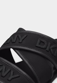 Gros plan sur les lanières d'une sandale noire avec l'inscription "DKNY" en relief et un tissu texturé sur un fond clair.