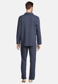 Mann steht barfuß mit dem Rücken zu uns, trägt navyblaue Pyjamas mit vertikalen hellblauen Streifen, langärmlig und mit langen Hosen.