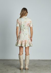 Beige Kurzarmkleid mit floralen Stickereien, Rüschensaum und gebundenem Taillenbund, kombiniert mit hohen beigen Cowboy-Stiefeln.