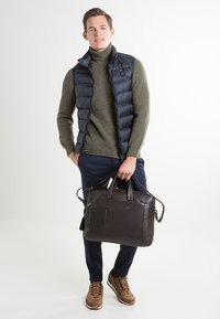 Gilet puffer blu sopra un maglione oliva, pantaloni blu, borsa in pelle marrone con manici superiori, sneakers marroni con lacci blu, sfondo semplice.