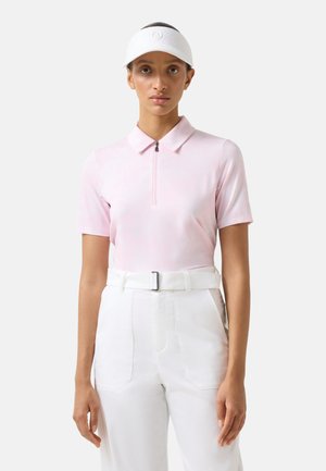 Vrouw draagt een lichtroze polo met korte mouwen en rits, witte broek met hoge taille en riem, en een witte zonnekap, staand tegen een effen achtergrond.