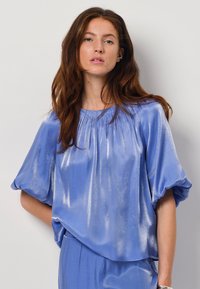 Blauwe satijnen blouse met een ontspannen pasvorm, korte pofmouwen en gathered halslijn. De stof heeft een subtiele glans en een lichte textuur.