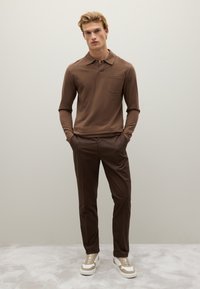 Braunes Langarm-Polohemd mit Tasche, kombiniert mit dunkelbraunen Hosen und weißen Sneakers mit beigen Akzenten, vor einem neutralen Hintergrund.