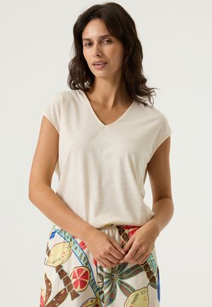 Beiges T-Shirt mit V-Ausschnitt und kurzen Ärmeln aus weichem Stoff, kombiniert mit gemusterten, bunten Hosen mit tropischem Design.