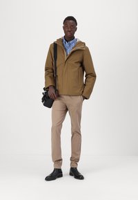 Giacca impermeabile marrone con cappuccio, rifinitura grigia e tasche con cerniera, abbinata a pantaloni beige e stivali neri, in piedi su uno sfondo bianco.