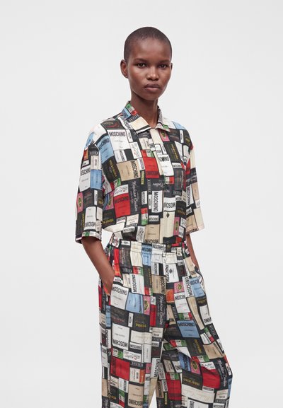 Personne debout portant une chemise à manches courtes et un pantalon assortis avec un imprimé coloré de l’étiquette Moschino, mains dans les poches, fond gris neutre.